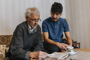 Male caregiver sitting with senior man playing Sudoku at home - MASF37642