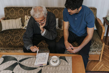 High angle view of senior man playing Soduko puzzle while sitting with male caregiver at home - MASF37643