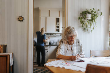 Senior woman doing puzzle in book while sitting at dining table in home - MASF37658