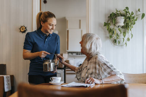 Glückliche ältere Frau im Gespräch mit einer weiblichen Pflegekraft, die eine Teekanne zu Hause hält - MASF37666