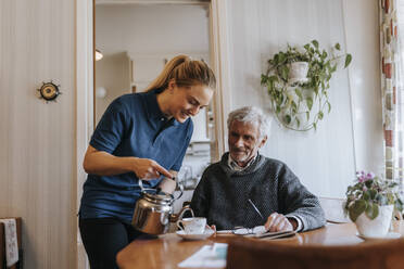 Lächelnde Pflegehelferin serviert Tee für einen älteren Mann, der zu Hause sitzt - MASF37674