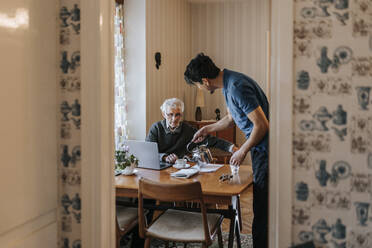 Side view of male healthcare worker serving tea to senior man at home - MASF37684