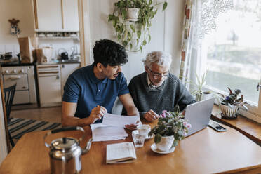 Male caregiver discussing medical reports with senior man using laptop at home - MASF37691