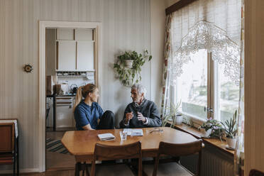 Älterer Mann diskutiert mit einer weiblichen Betreuerin, während er am Esstisch in seinem Haus sitzt - MASF37694