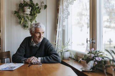 Senior man looking through window while sitting at dining table in home - MASF37696
