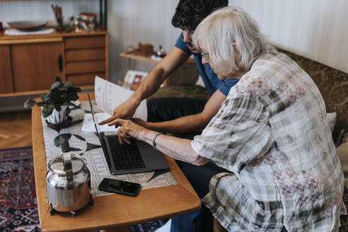 Ältere Frau diskutiert am Laptop, während sie mit einem männlichen Betreuer zu Hause sitzt - MASF37698