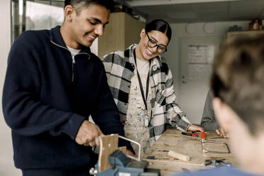 Happy female teacher standing with male student cutting wood during carpentry class - MASF37709