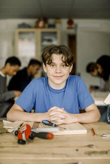 Porträt eines lächelnden männlichen Teenagers mit verschränkten Händen am Tisch während des Schreinerunterrichts - MASF37717