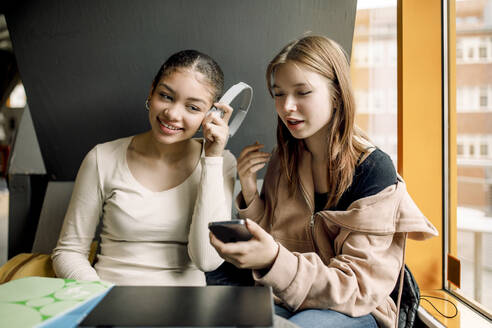Lächelndes Mädchen, das mit einer Freundin, die ein Smartphone hält, in der Schule sitzt und Musik hört - MASF37726