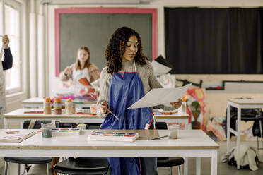 Teenager-Schülerin, die während des Kunstunterrichts in der High School ein Gemälde am Schreibtisch betrachtet - MASF37745