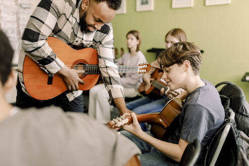 Junger männlicher Lehrer, der einem Teenager beim Gitarrenspiel im Musikunterricht an einer High School hilft - MASF37756