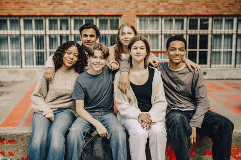 Portrait of smiling male and female students sitting with arms around in high school campus - MASF37778