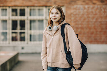 Porträt einer lächelnden Teenager-Schülerin mit Rucksack und Kopfhörern auf dem Campus einer High School - MASF37781