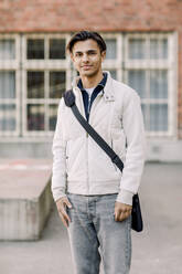 Porträt eines lächelnden männlichen Teenager-Schülers, der mit einer Umhängetasche auf dem Highschool-Campus steht - MASF37788