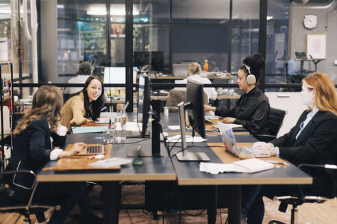 Happy young businesswoman talking to female colleague while working together at desk in office - MASF37797