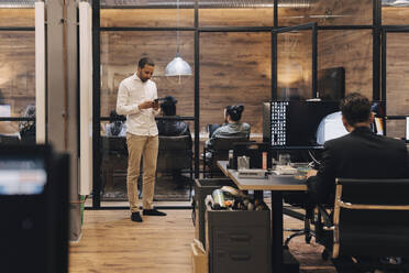 Businessman using smart phone near colleague using computer at desk in office - MASF37840