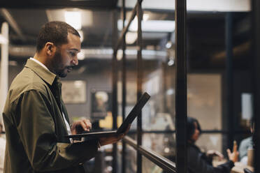 Side view of dedicated businessman using laptop while standing in illuminated office - MASF37848