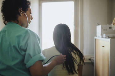 Rear view of mature female doctor examining young woman with stethoscope in clinic - MASF37872