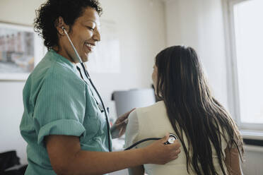 Side view of smiling mature female doctor examining young woman with stethoscope in clinic - MASF37873