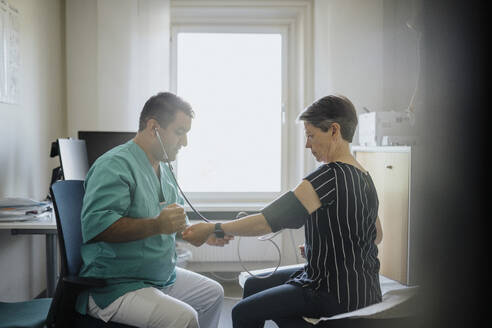 Male physician checking blood pressure of senior woman while sitting in clinic - MASF37884