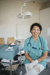 Portrait of happy female doctor wearing medical scrubs sitting in medical examination room - MASF37919