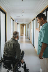 Male doctor showing way to male patient sitting in wheelchair at hospital corridor - MASF37921