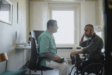 Side view of mature male doctor discussing with patient sitting in wheelchair at clinic - MASF37929