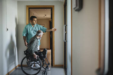 Doctor pointing male patient in wheelchair towards entrance of clinic - MASF37930