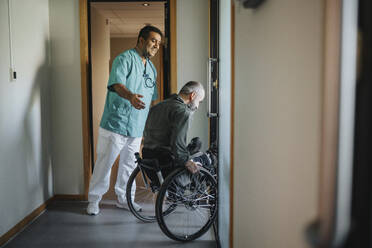 Happy doctor assisting patient in wheelchair at corridor of clinic - MASF37931
