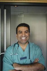 Porträt eines fröhlichen reifen Arztes, der mit verschränkten Armen vor einer Tür im Krankenhaus steht - MASF37932