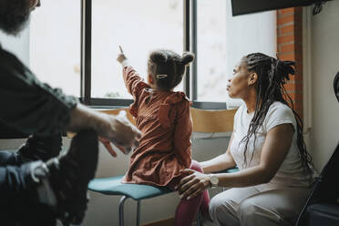 Mother with daughter pointing at window while waiting in hospital - MASF37940