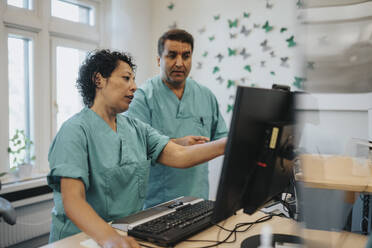 Male and female healthcare workers discussing over computer at reception desk in hospital - MASF37943