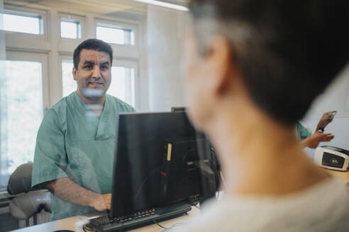 Male receptionist talking with patient through glass screen at reception desk in hospital - MASF37948
