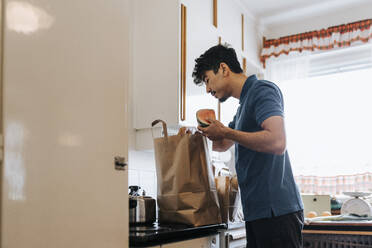 Seitenansicht eines männlichen Pflegehelfers beim Auspacken von Lebensmitteln aus einer Tüte in der Küche zu Hause - MASF37950