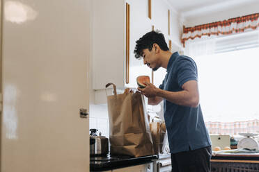 Side view of male care assistant unpacking groceries from bag in kitchen at home - MASF37950