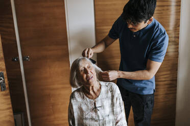 Männlicher Pflegehelfer kämmt das Haar einer älteren Frau zu Hause - MASF37971