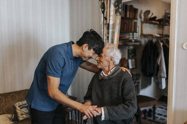 Lächelnder männlicher Pflegehelfer berührt die Stirn eines älteren Mannes zu Hause - MASF37978