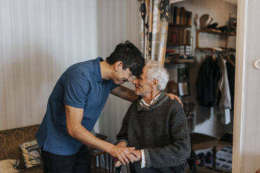 Smiling male care assistant touching forehead with senior man at home - MASF37978