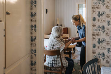 Lächelnde ältere Frau, die die Hände einer Mitarbeiterin im Gesundheitswesen hält, während sie zu Hause auf einem Stuhl sitzt - MASF37984