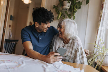 Happy male caregiver with senior woman laughing at home - MASF37993