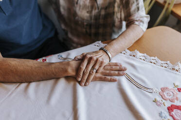Ältere Frau hält die Hand eines männlichen Pflegers über einem Tischtuch zu Hause - MASF37995