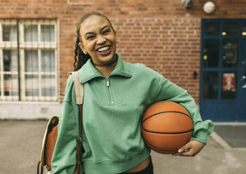 Porträt einer glücklichen jungen Frau, die einen Basketball unter dem Arm hält, während sie vor einem Gebäude steht - MASF37997