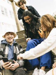 Low angle view of male and female friends sharing smart phone while sitting on steps - MASF38001