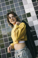 Porträt einer lächelnden jungen Frau, die eine Jacke hält, während sie an einer gekachelten Wand steht - MASF38020
