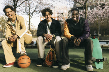 Glückliche multirassische männliche Freunde, die lachend auf einer Bank sitzen, während eines sonnigen Tages - MASF38033