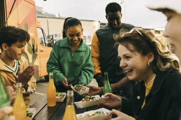 Happy male and female friends enjoying snacks with drinks near food truck - MASF38039