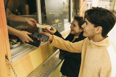 Smiling young man doing online payment while buying food from concession stand - MASF38043