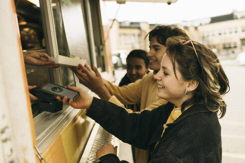 Smiling young woman doing online payment through smart phone while buying food from concession stand - MASF38044