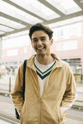 Happy young man with hands in pockets while standing at train station - MASF38050