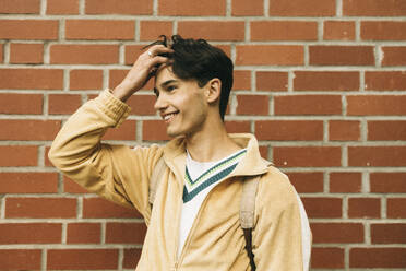 Smiling young man with hand in hair while standing against brick wall - MASF38055